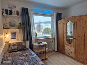 A cozy bedroom with a bed, a small table, a wardrobe, and a window with a view of trees and a building.
