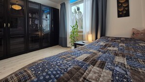 A bedroom with a large bed, dark wooden wardrobe, a window with curtains, a bedside table with a lamp, and a wall with a framed picture.