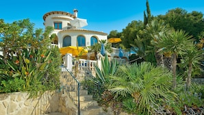 Uma vila em estilo mediterrâneo com uma escadaria de pedra que leva a uma varanda, rodeada por plantas tropicais exuberantes e um céu azul límpido.