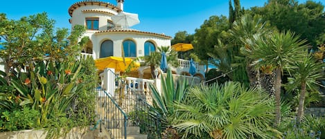 A Mediterranean-style villa with a stone staircase leading to a balcony, surrounded by lush tropical plants and a clear blue sky.