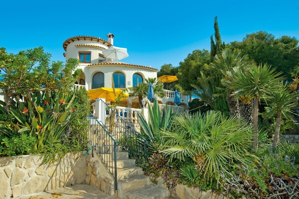 A Mediterranean-style villa with a stone staircase leading to a balcony, surrounded by lush tropical plants and a clear blue sky.