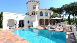 Uma área de piscina com piscina azul cristalina, espreguiçadeiras brancas e um edifício com arcos e uma torre.