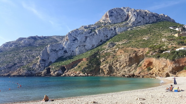 Litoral rochoso com águas turquesa cristalinas, uma praia de seixos e uma montanha ao fundo.