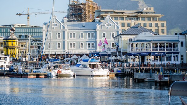 Una marina con barcos amarrados, un edificio con torre de reloj y grúas de construcción al fondo.