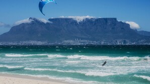 Una playa con una persona practicando windsurf, cielo despejado y una montaña al fondo.