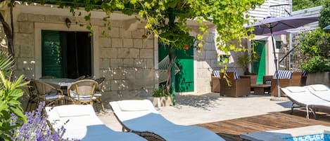 A wooden deck with lounge chairs, a pool, and a stone building with green shutters.
