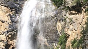 Uma cascata desce por um penhasco rochoso, cercada por vegetação.