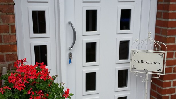 Eine weiße Tür mit einem 'Willkommen'-Schild, einem Schlüssel im Schloss und einem Blumentopf mit roten Blüten.