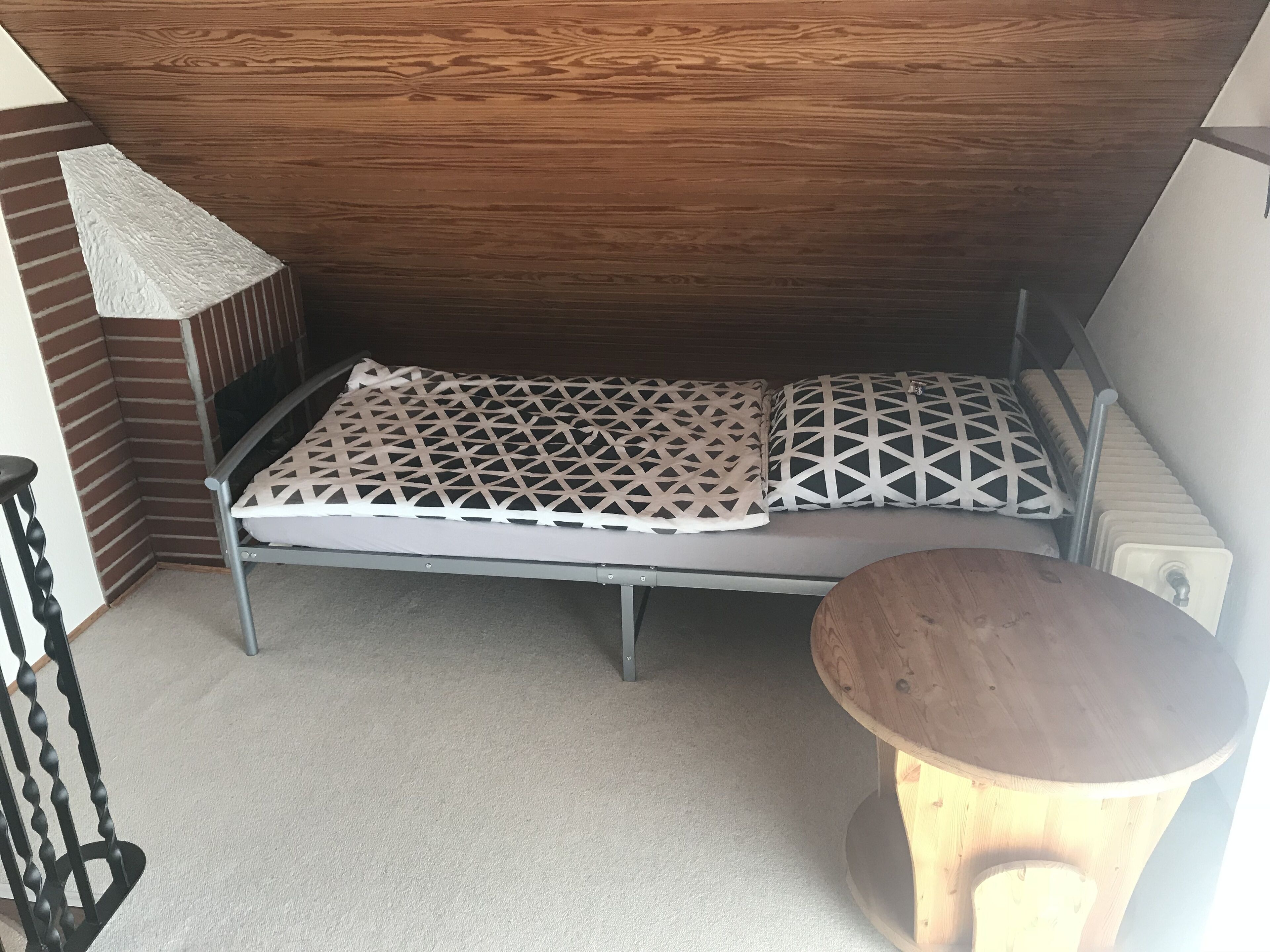 A compact bed with a geometric-patterned mattress, a small round wooden table, and a radiator on the wall.