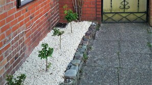 Ein Ziegelhaus mit Fenster, ein kleiner Garten mit weißen Kieselsteinen und Pflanzen, und ein gelbes Tor mit schmiedeeisernen Verzierungen.