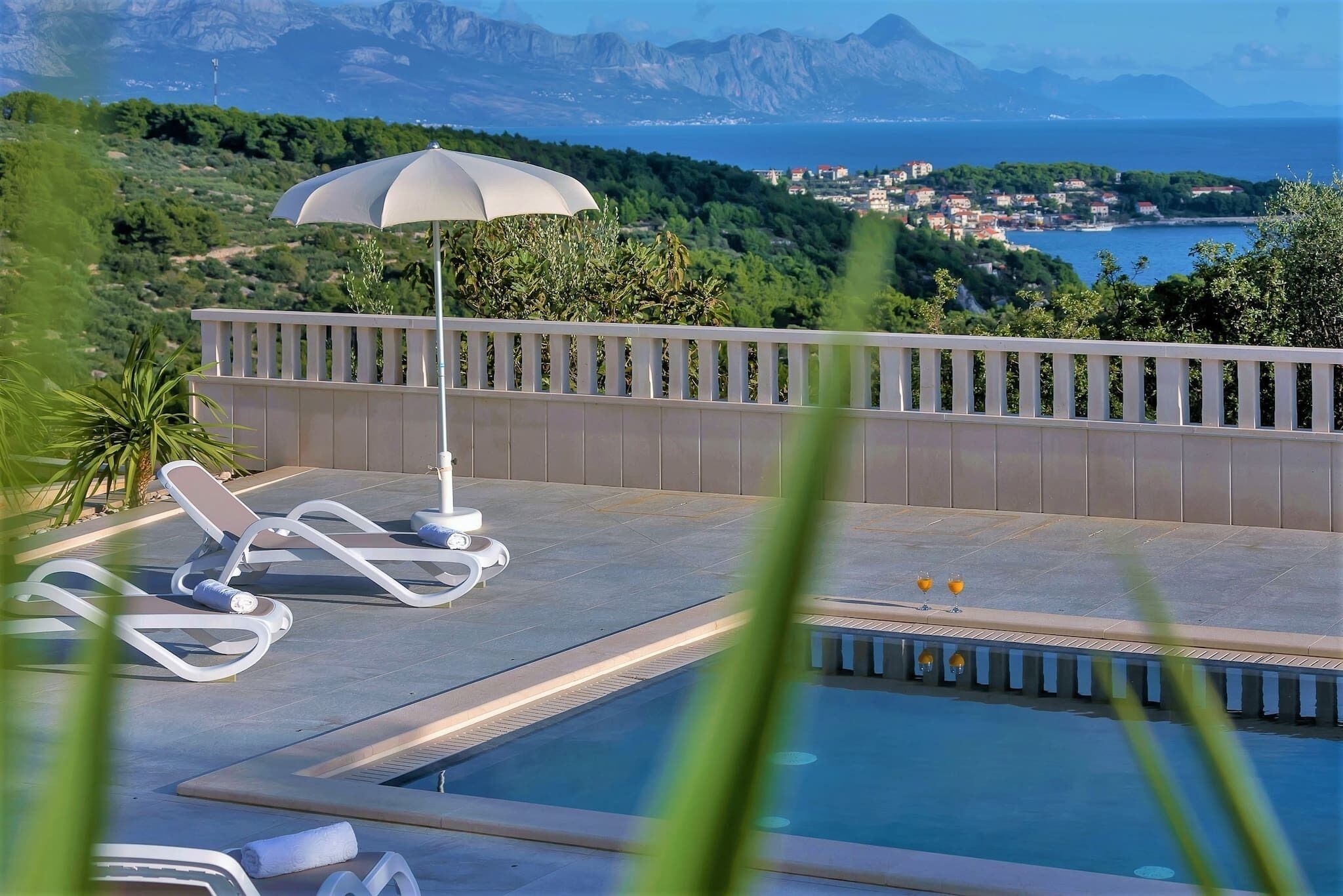 Ein Poolbereich mit Liegestühlen und Sonnenschirm, mit Blick auf eine Küstenstadt und Berge.