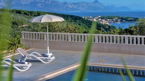 Un espace aménagé au bord de la piscine, avec des chaises longues et un parasol, offrant une vue sur une ville côtière et les montagnes.