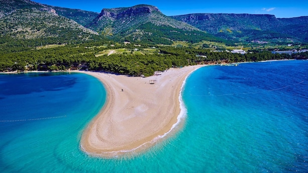Vue aérienne d’une plage aux eaux turquoise et limpides, bordée d’un rivage sablonneux, avec une chaîne de montagnes en arrière-plan.