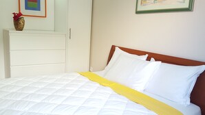 A neatly made bed with white and yellow bedding, a wooden headboard, and a white dresser with a vase of red flowers. - Ginkgo 3 - Holiday apartment for 3 people, close to the sea (Banjole)