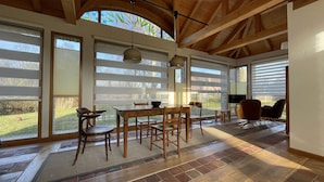 A dining area with a wooden table and chairs, large windows with blinds, and a view of the outdoors.