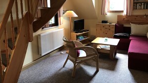 Una habitación acogedora con una escalera de madera, un televisor, una estantería, una ventana con cortinas y una zona de estar confortable.