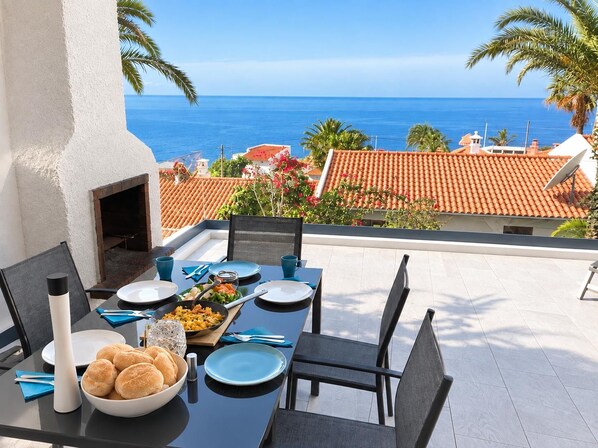 Mesa de comedor para cuatro personas con vista al mar y un tejado de tejas rojas al fondo.