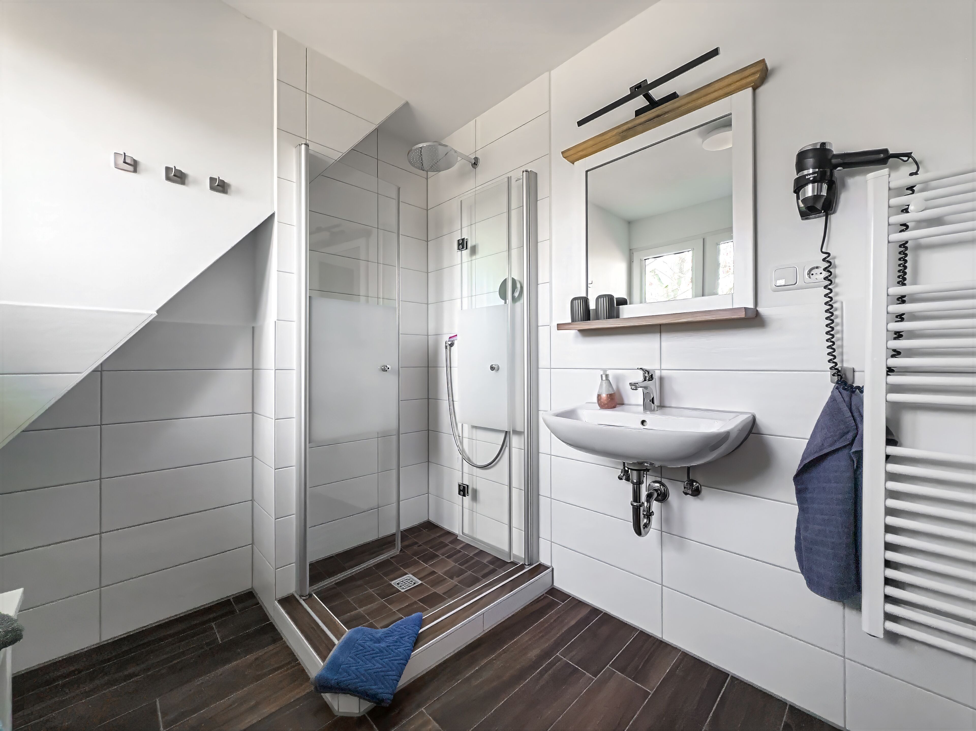 Un baño moderno con cabina de ducha de vidrio, un lavabo blanco, un perchero y un estante de madera.