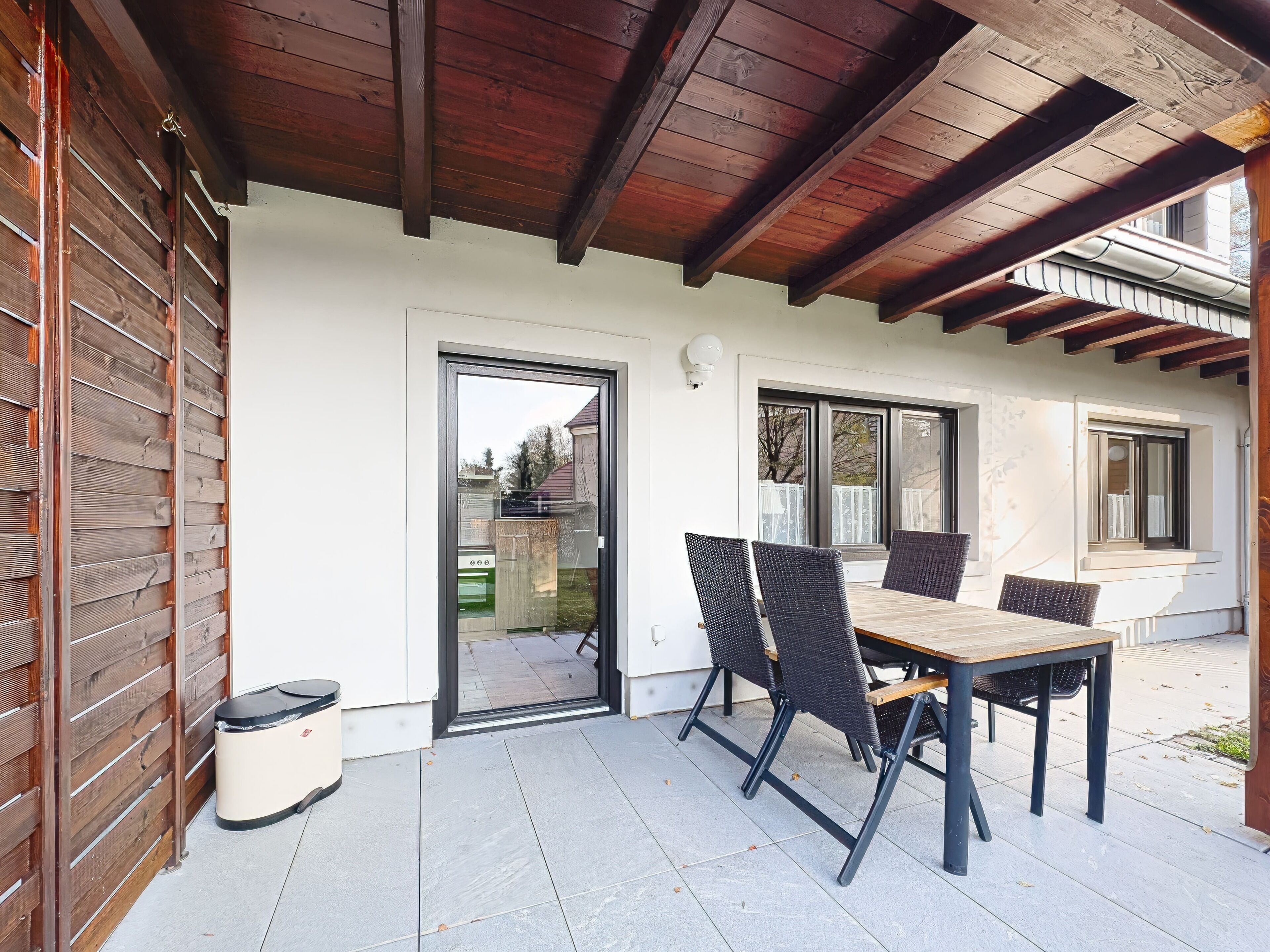 A covered patio with a table and chairs, a door leading to the interior, and a view of the outside.