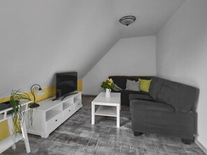 A living room with a TV on a white stand, a grey sofa, a white coffee table, and a ceiling light fixture.