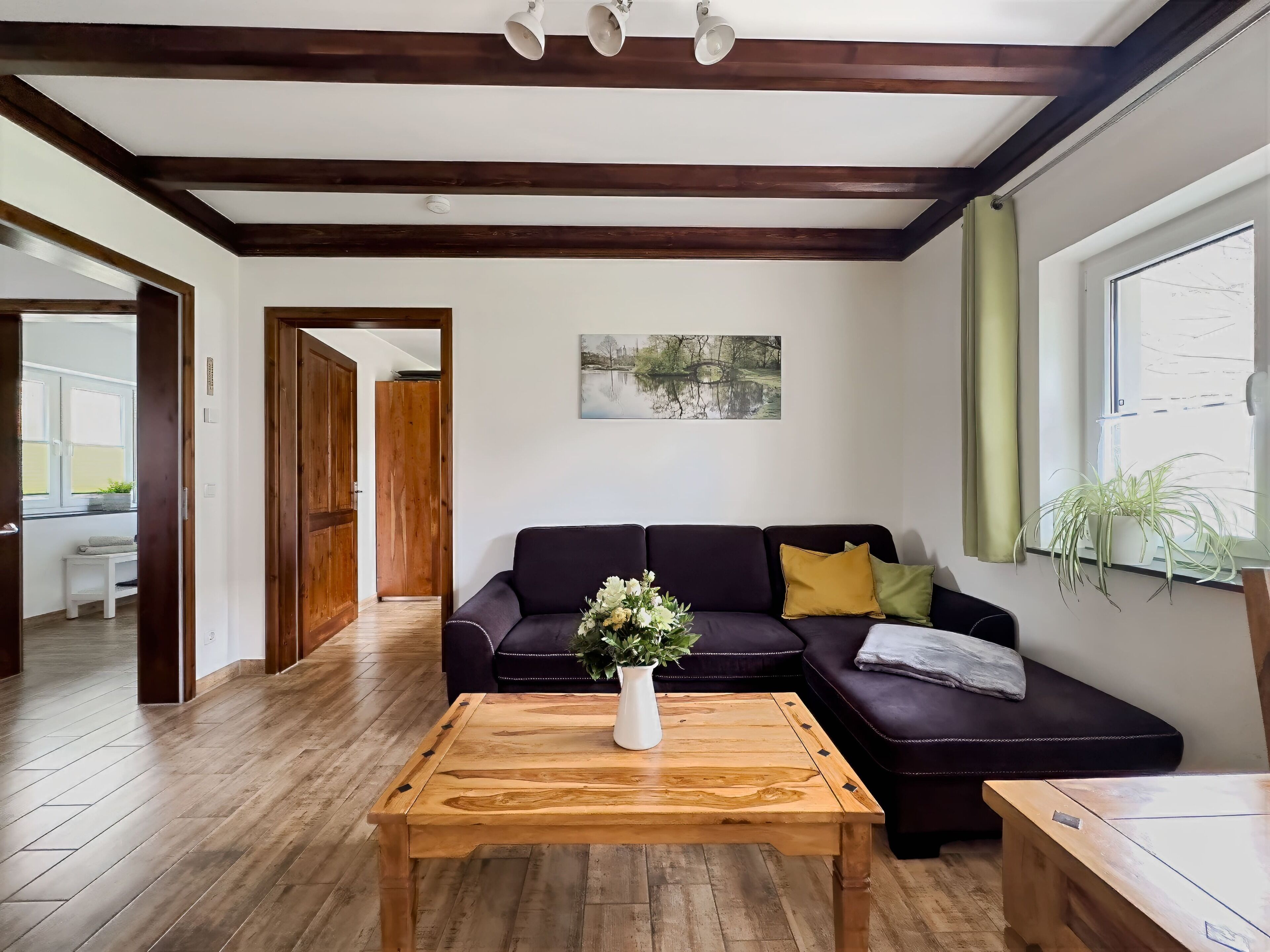 Een woonkamer met een donkere bank, een houten salontafel, een schilderij aan de wand en een raam met gordijnen.