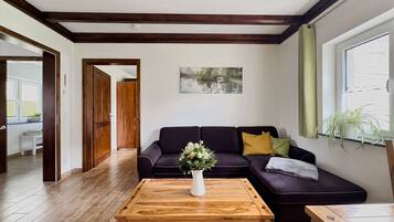Een woonkamer met een donkere bank, een houten salontafel, een schilderij aan de wand en een raam met gordijnen.