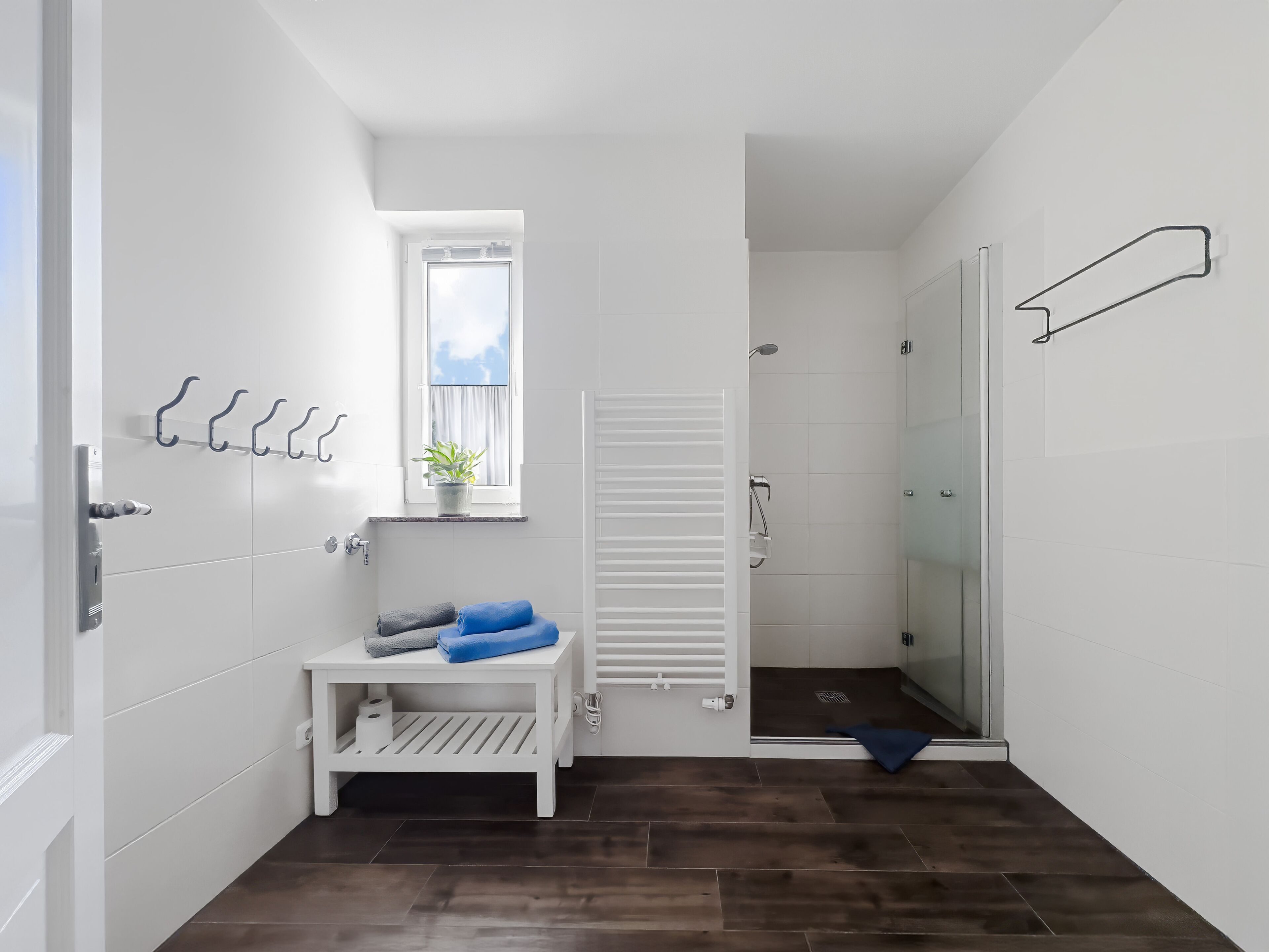 A modern bathroom with white tiles, a shower, a sink with a towel rack, and a small window with a plant.