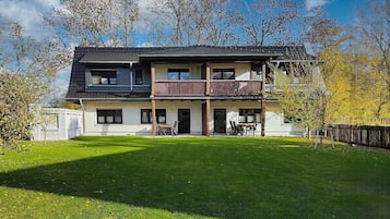 Una casa a due piani con un balcone, un prato davanti e alberi sullo sfondo.