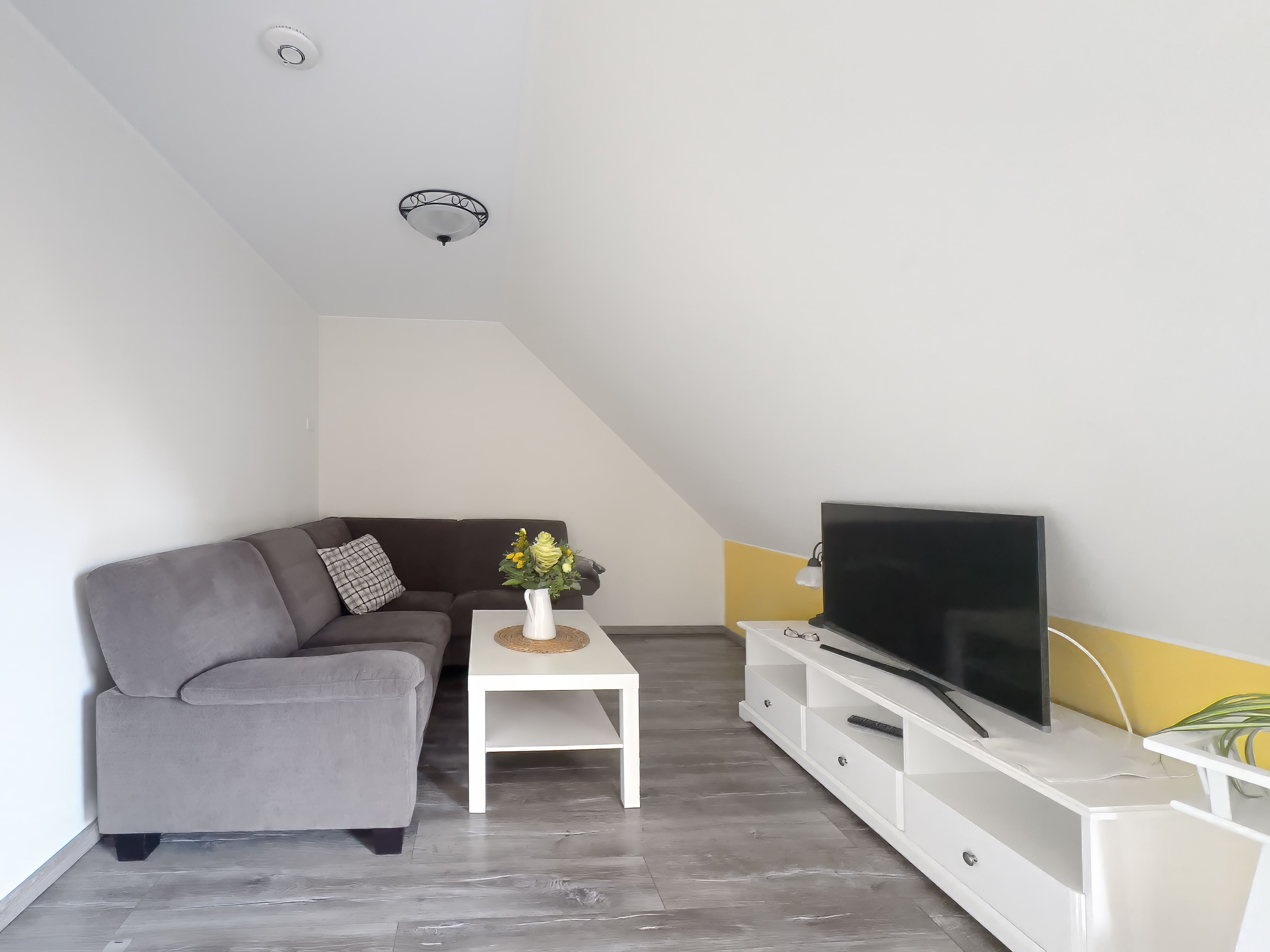 En moderne stue med en grå sofa, et hvidt sofabord med en vase, et fladskærms-tv på et hvidt tv-bord, og en loftlampe.