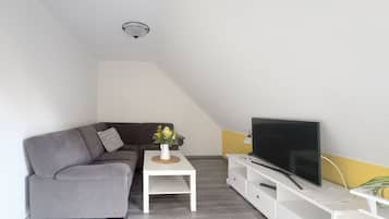 En moderne stue med en grå sofa, et hvidt sofabord med en vase, et fladskærms-tv på et hvidt tv-bord, og en loftlampe.