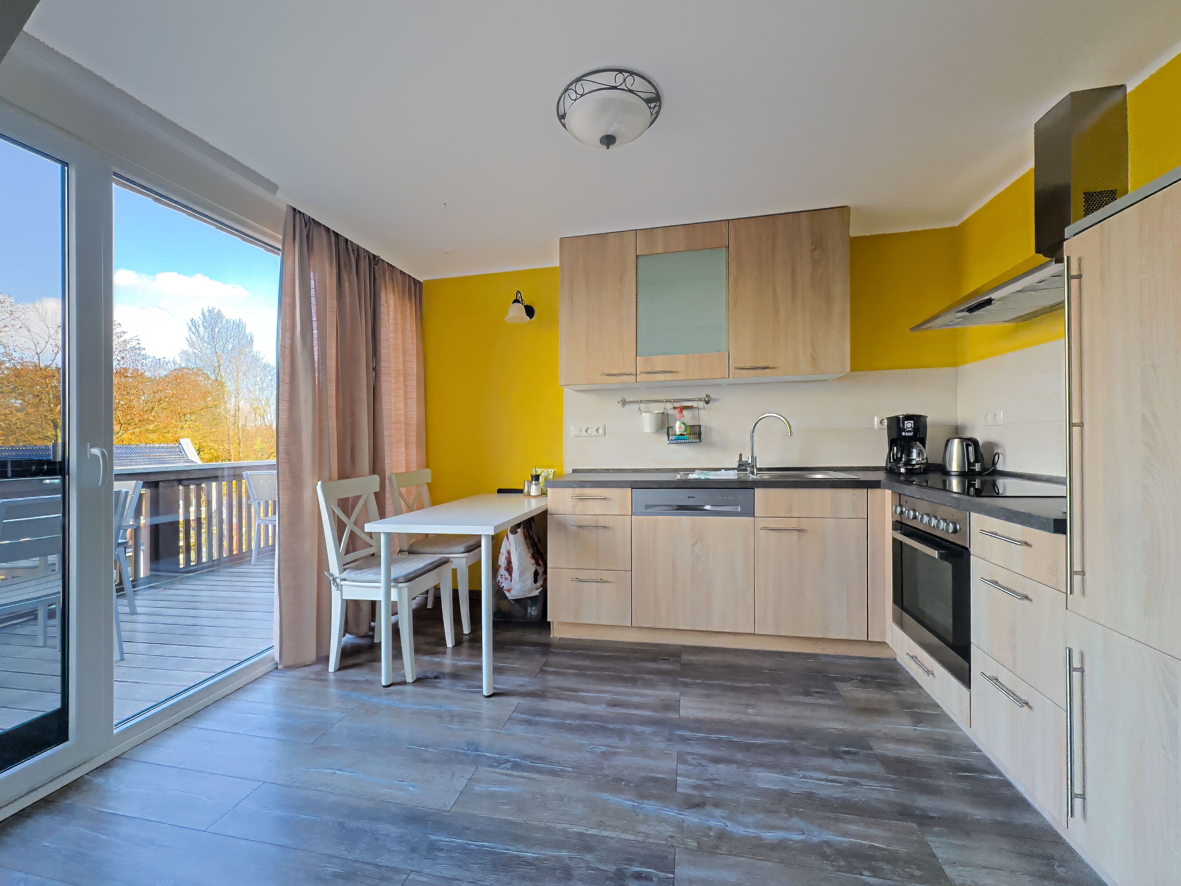 Una cocina moderna con armarios de madera, una mesa de comedor con sillas, un lavavajillas y un ventanal con vistas a árboles.