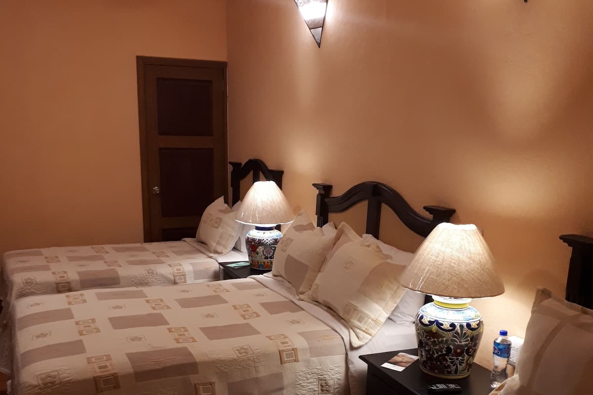 Ein Hotelzimmer mit zwei Betten, jedes mit einer gemusterten Tagesdecke und zwei Kissen. Zwei Nachttische sind vorhanden, jeder mit einer Lampe und einer Fernbedienung. Auf einem Tisch steht eine Wasserflasche.