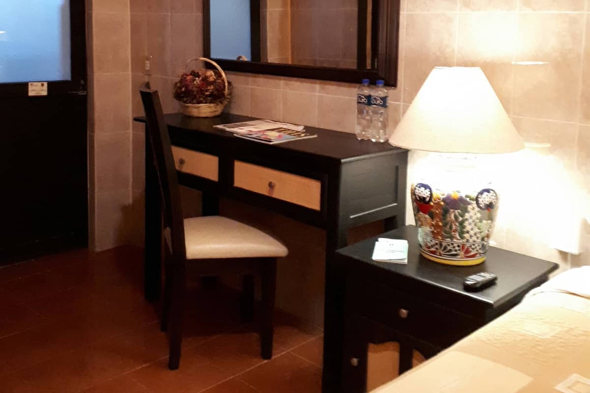Ein Hotelzimmer mit Schreibtisch, Stuhl und Spiegel. Auf einem Beistelltisch steht eine Lampe, auf dem Schreibtisch befinden sich eine Blumenkorb und zwei Wasserflaschen.