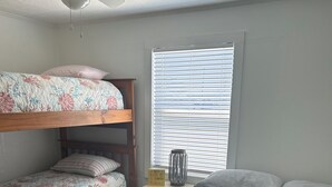 A bedroom with two bunk beds, a single bed, a ceiling fan, a window with blinds, and a small table with a candle and a sign. - Cozy beach cottage in Magnolia (Port Lavaca)