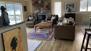 Un salon chaleureux avec une cheminée en pierre, des sièges confortables et une grande fenêtre donnant sur l’extérieur.