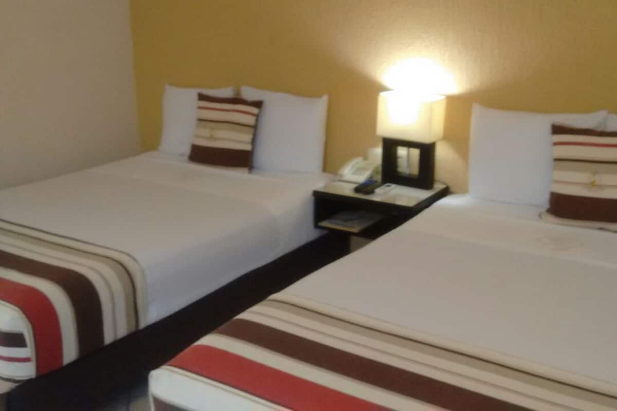 A hotel room with two beds, each with striped bedding and pillows. There is a nightstand between the beds with a lamp and a telephone.