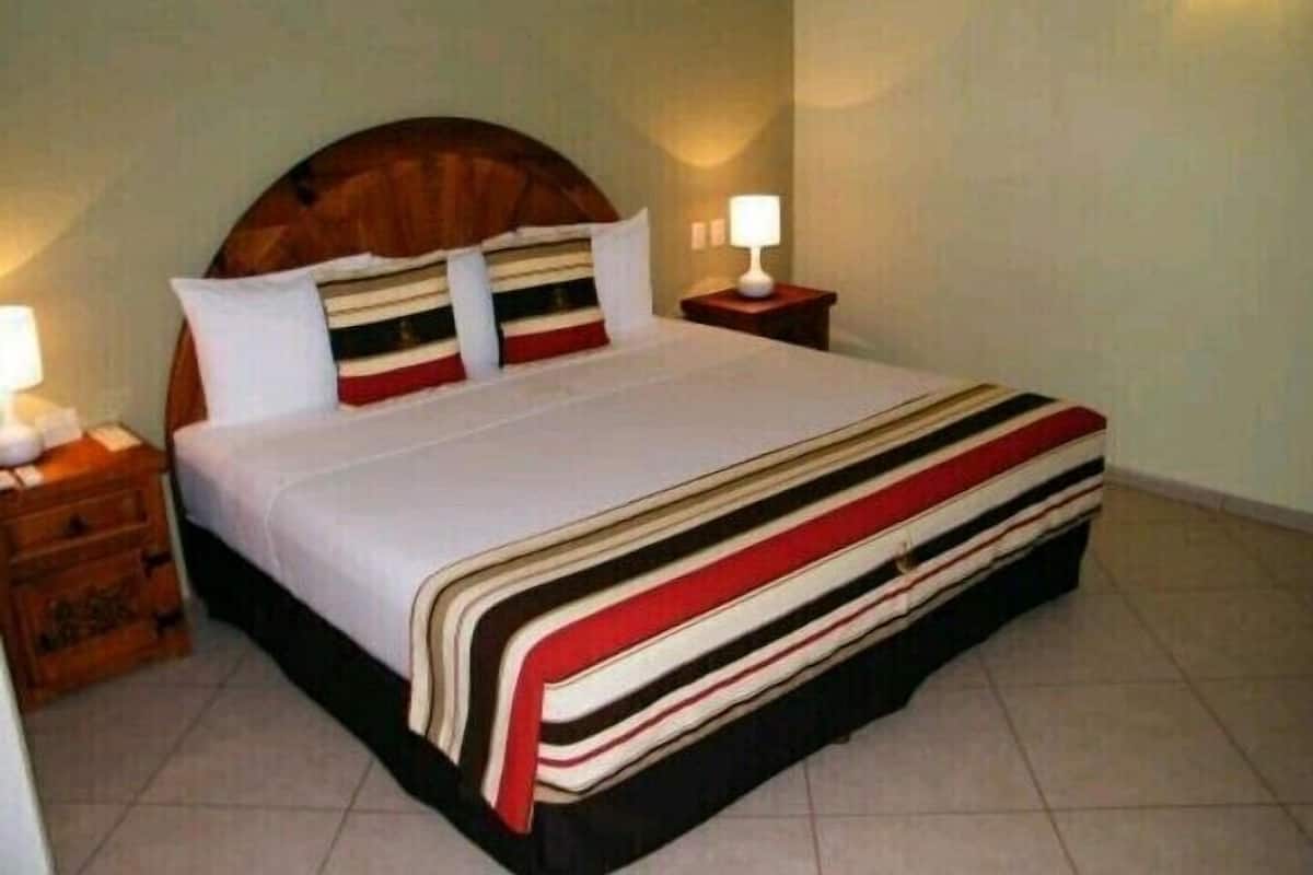 A neatly made bed with a striped bedspread and pillows, a wooden headboard, a nightstand with a lamp, and a wall-mounted light fixture.