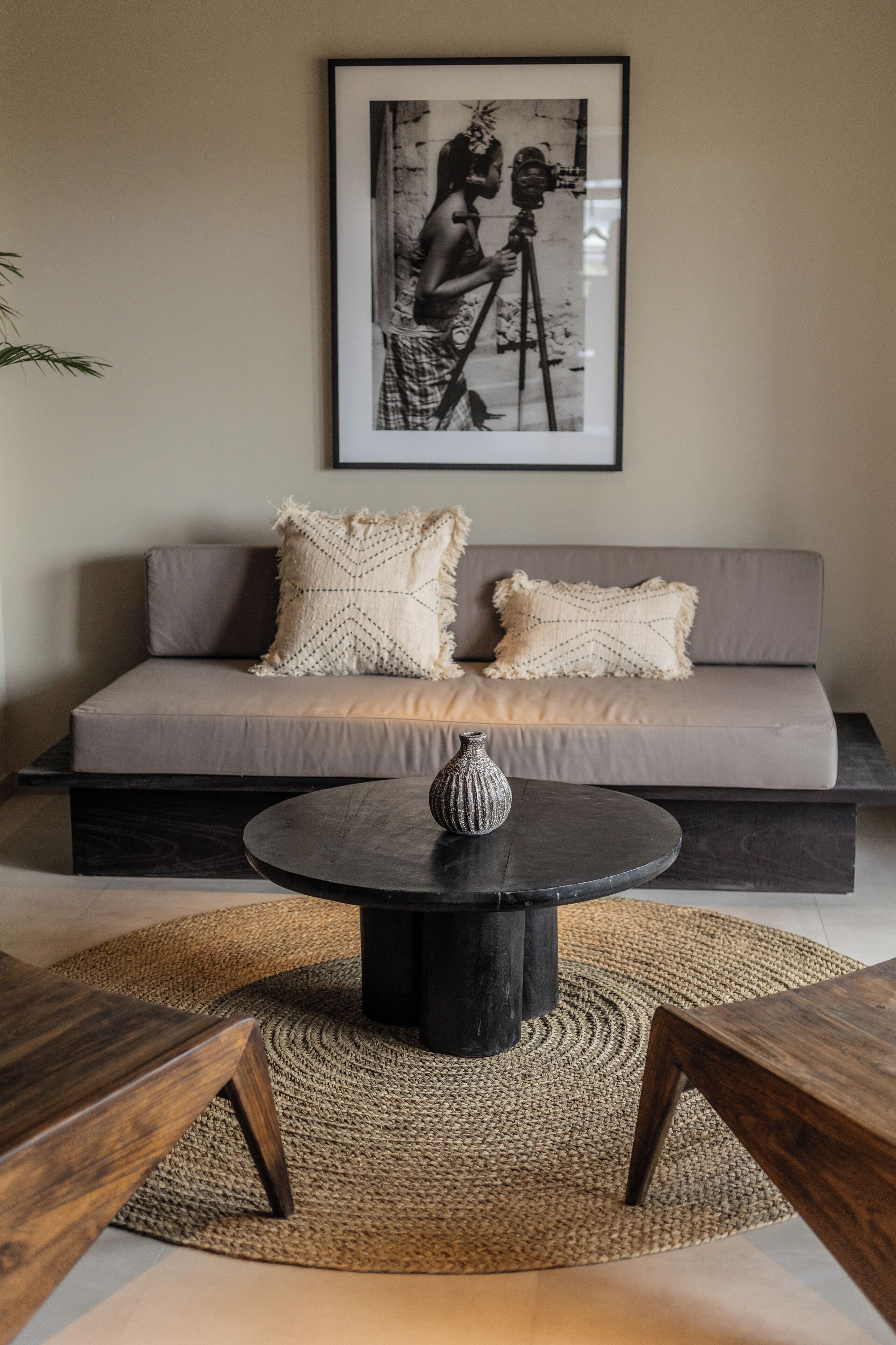 Een moderne woonkamer met een grijze bank, twee kussens met een patroon, een ronde zwarte salontafel met een vaas, en een zwart-wit foto in een lijst aan de muur.