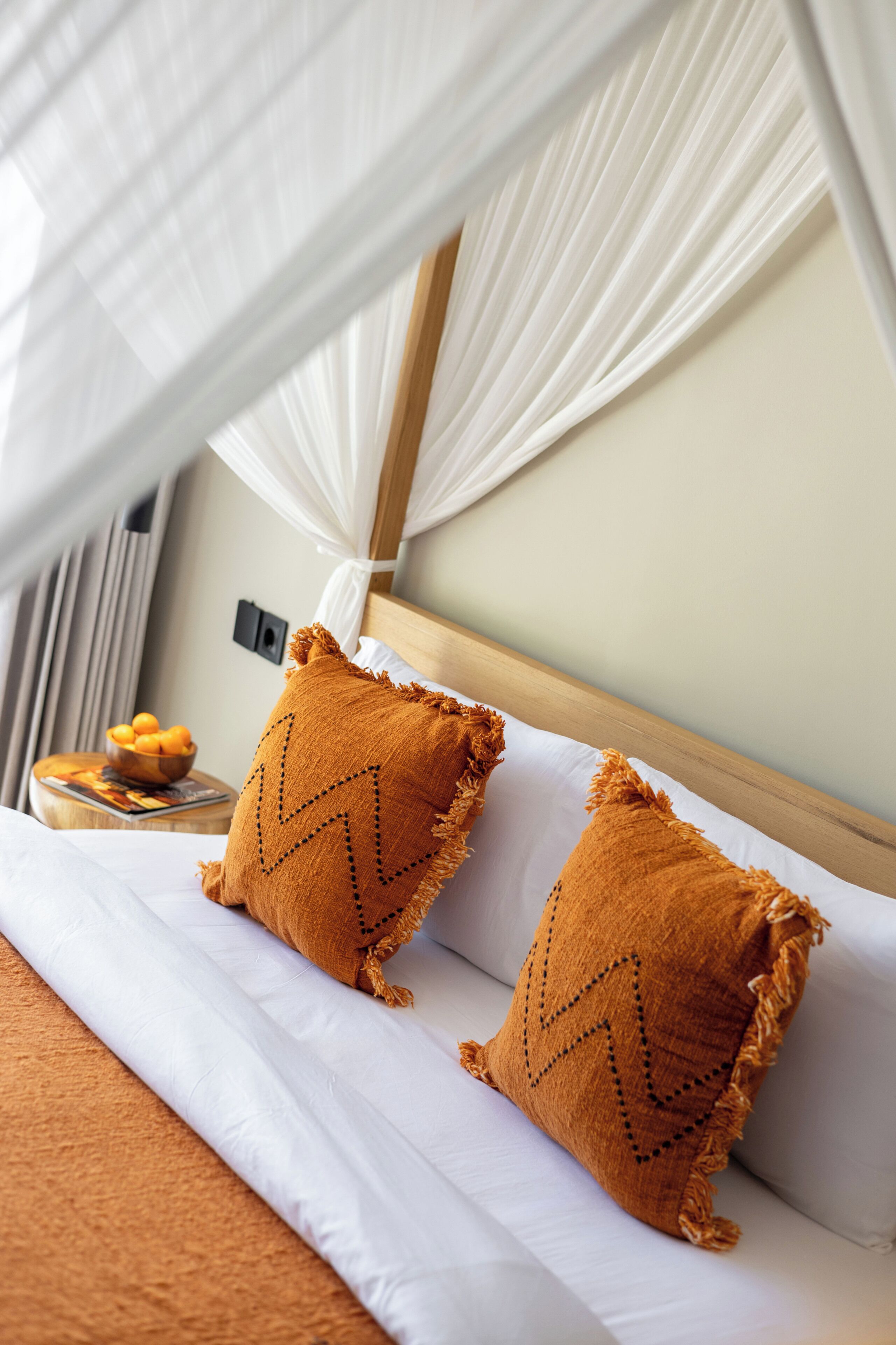 Hotelkamer met een bed, wit beddengoed, twee oranje kussens, een houten hoofdbord, een nachtkastje met een schaal fruit en een wit baldakijn.
