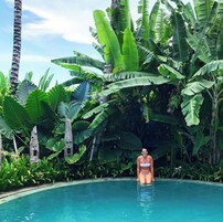 Mulher de biquíni parada em uma piscina, cercada por folhagem tropical exuberante.