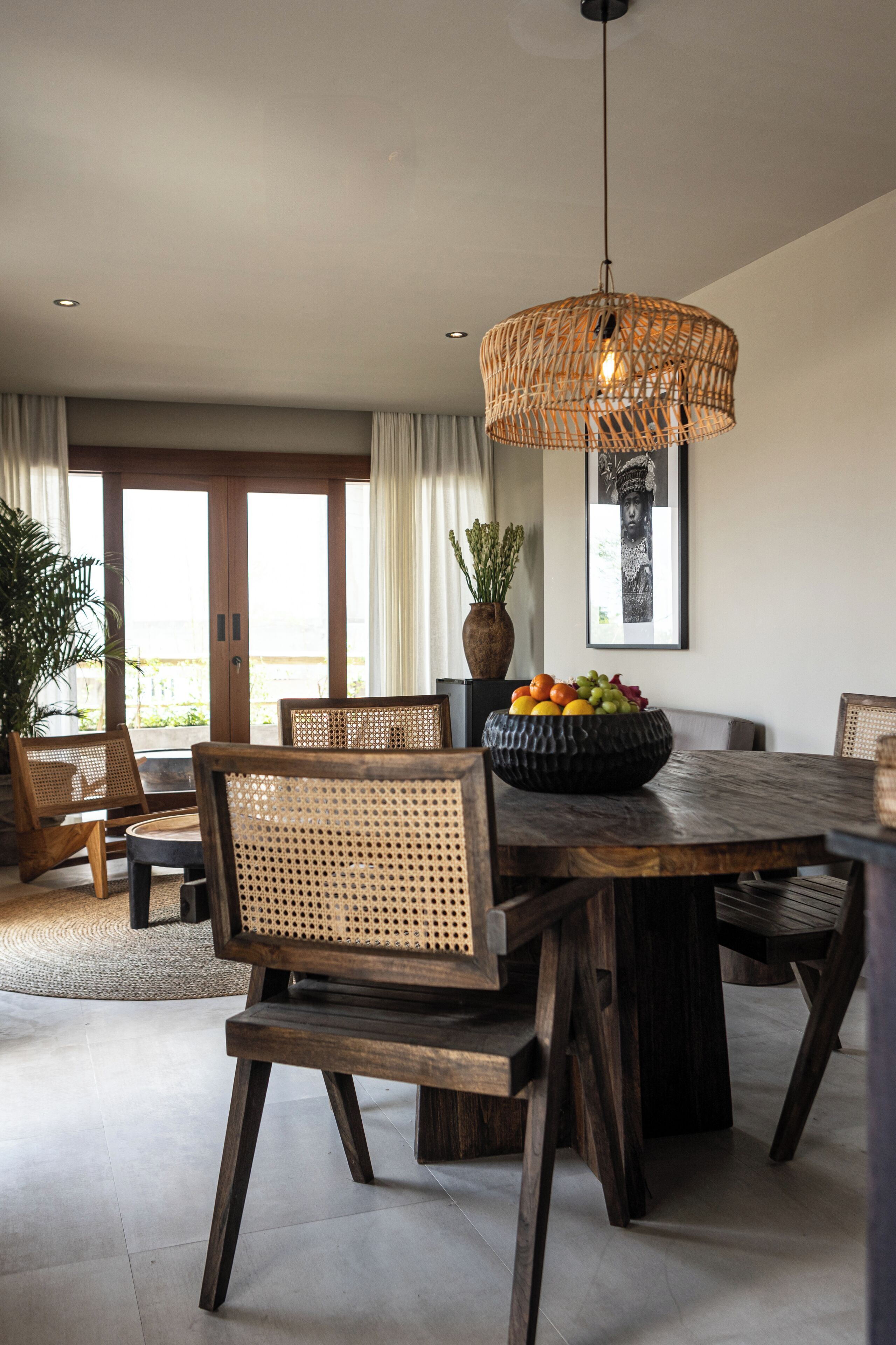 Un comedor con una mesa redonda de madera, sillas de mimbre, una lámpara colgante y un frutero sobre la mesa.