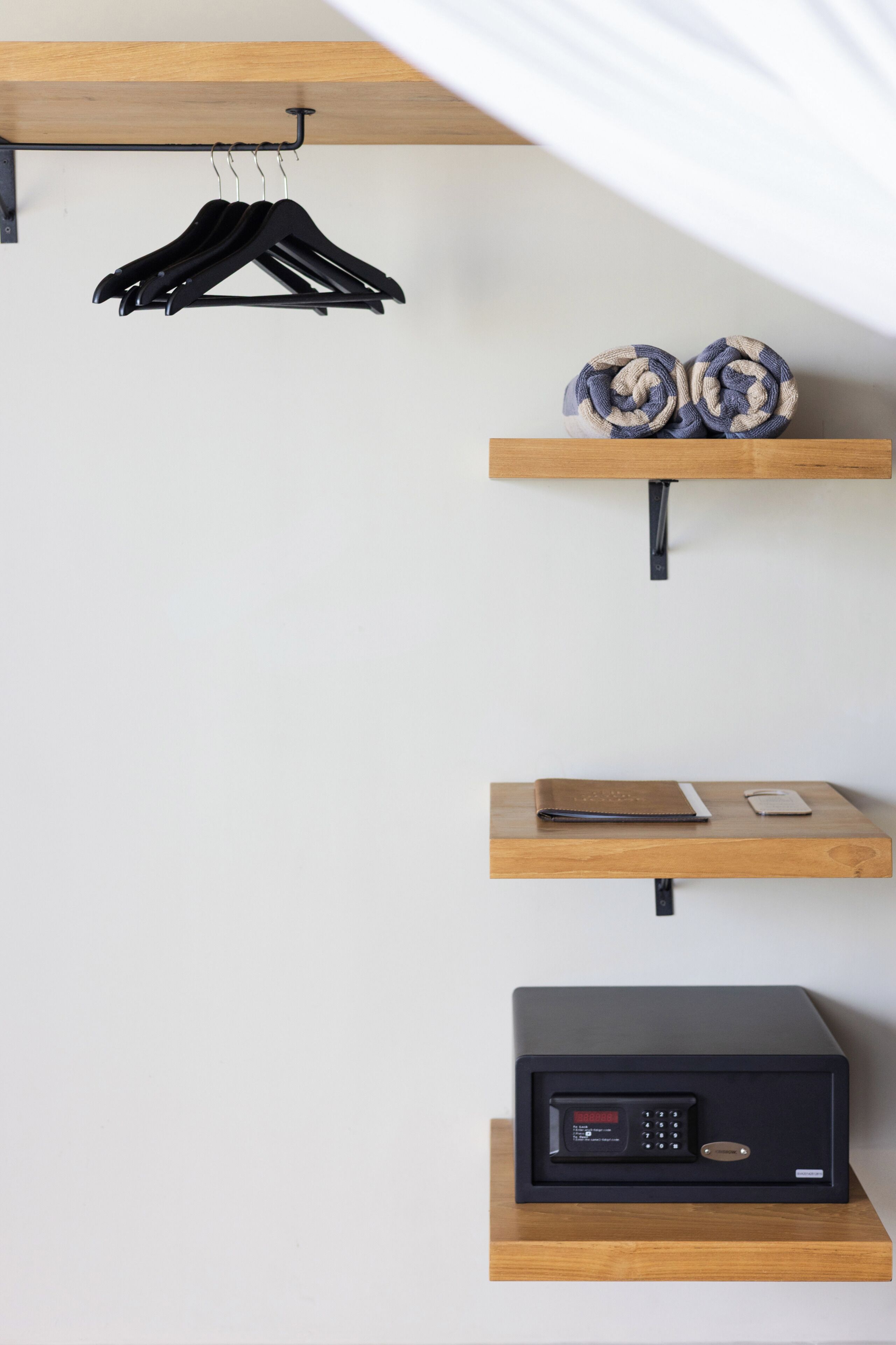 A room with a wooden shelf holding a black electronic device, a wooden shelf with rolled towels, and a wooden shelf with a black hanger.