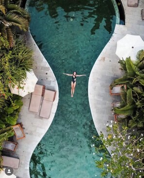 Una piscina con una persona flotando en el centro, rodeada de sillas de descanso y palmeras.
