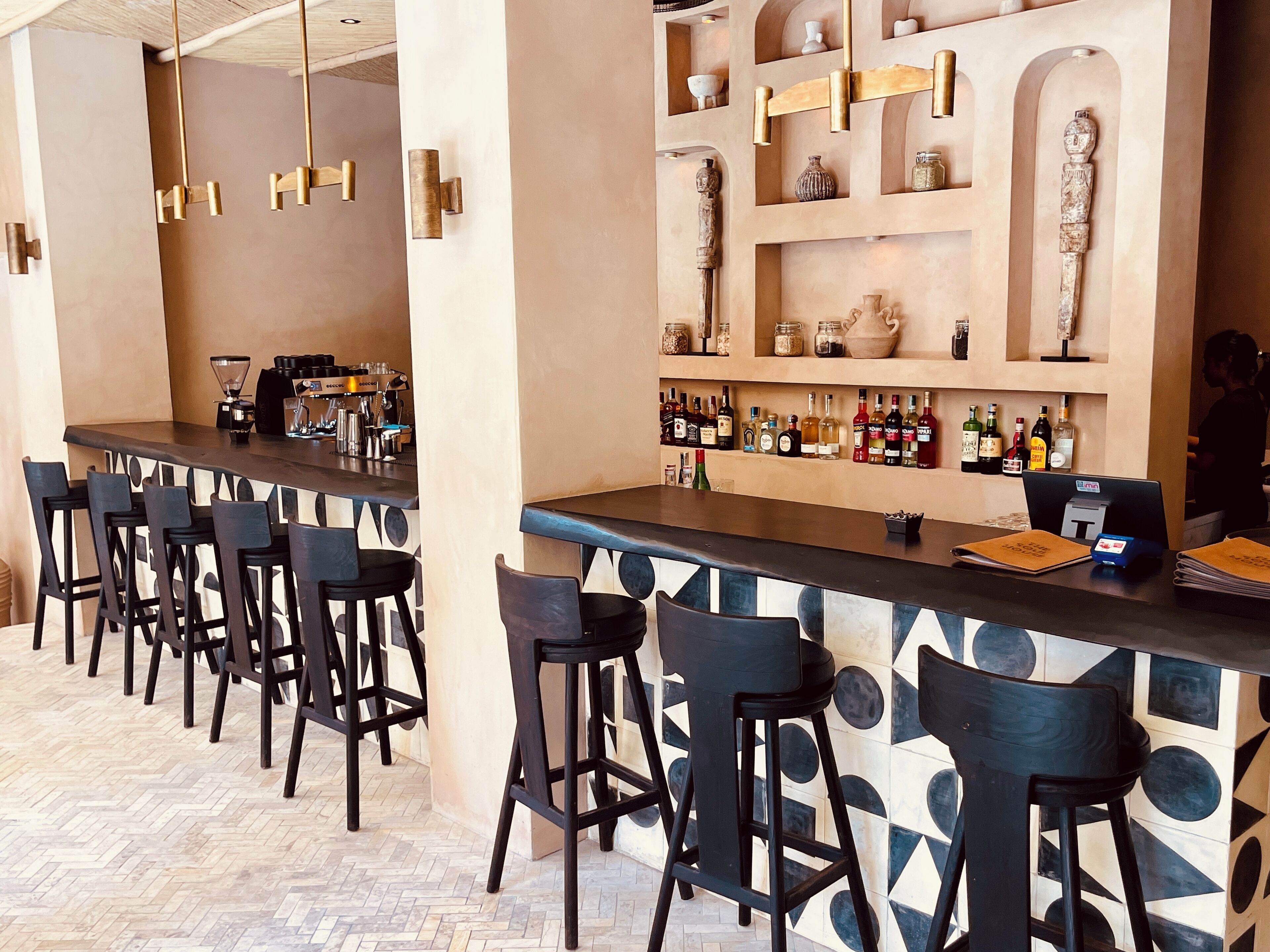 A bar area with a long counter, multiple stools, and shelves with bottles and decorative items.