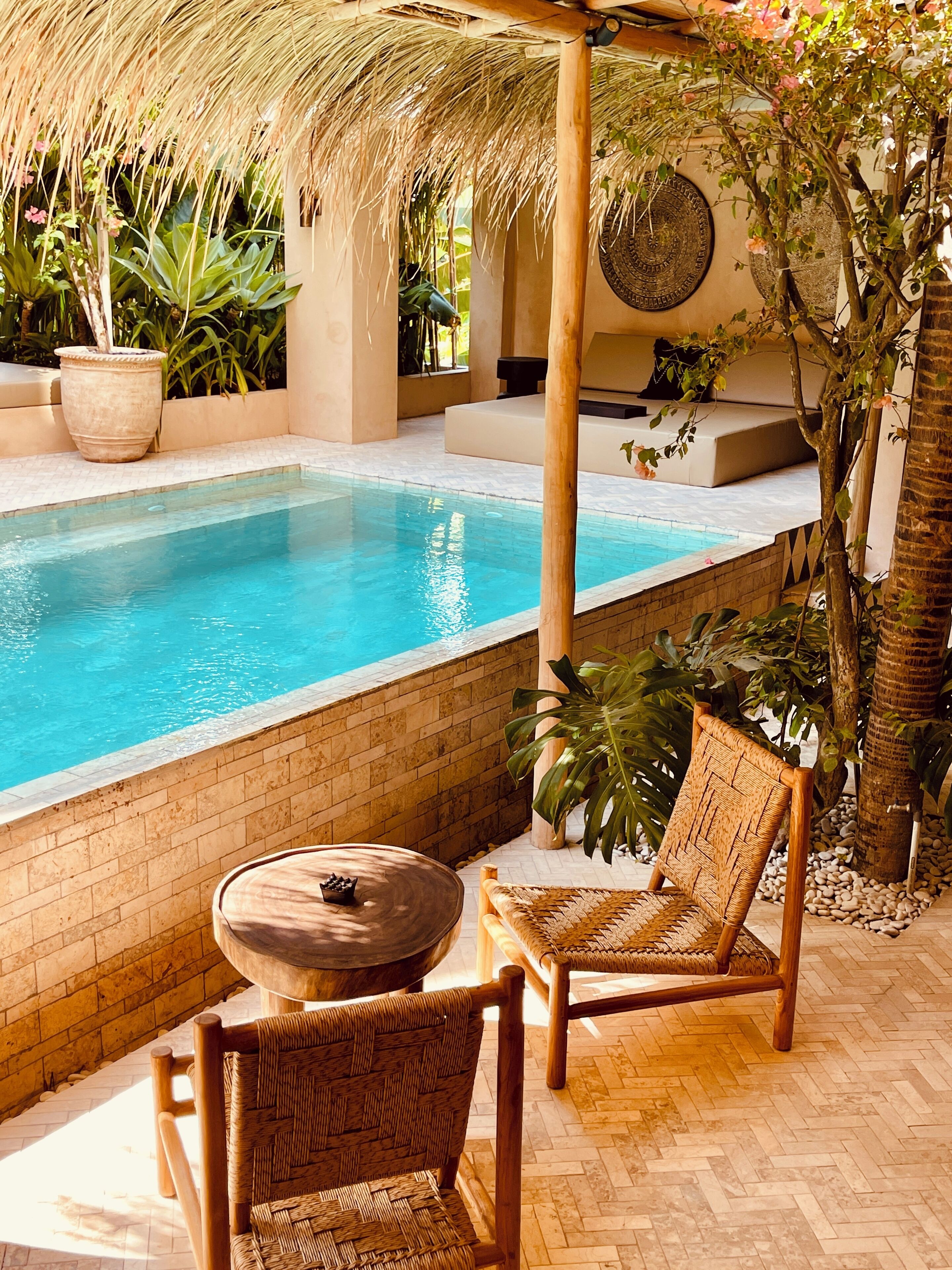 Uma área à beira da piscina com parede de tijolos, telhado de palha e móveis de vime.