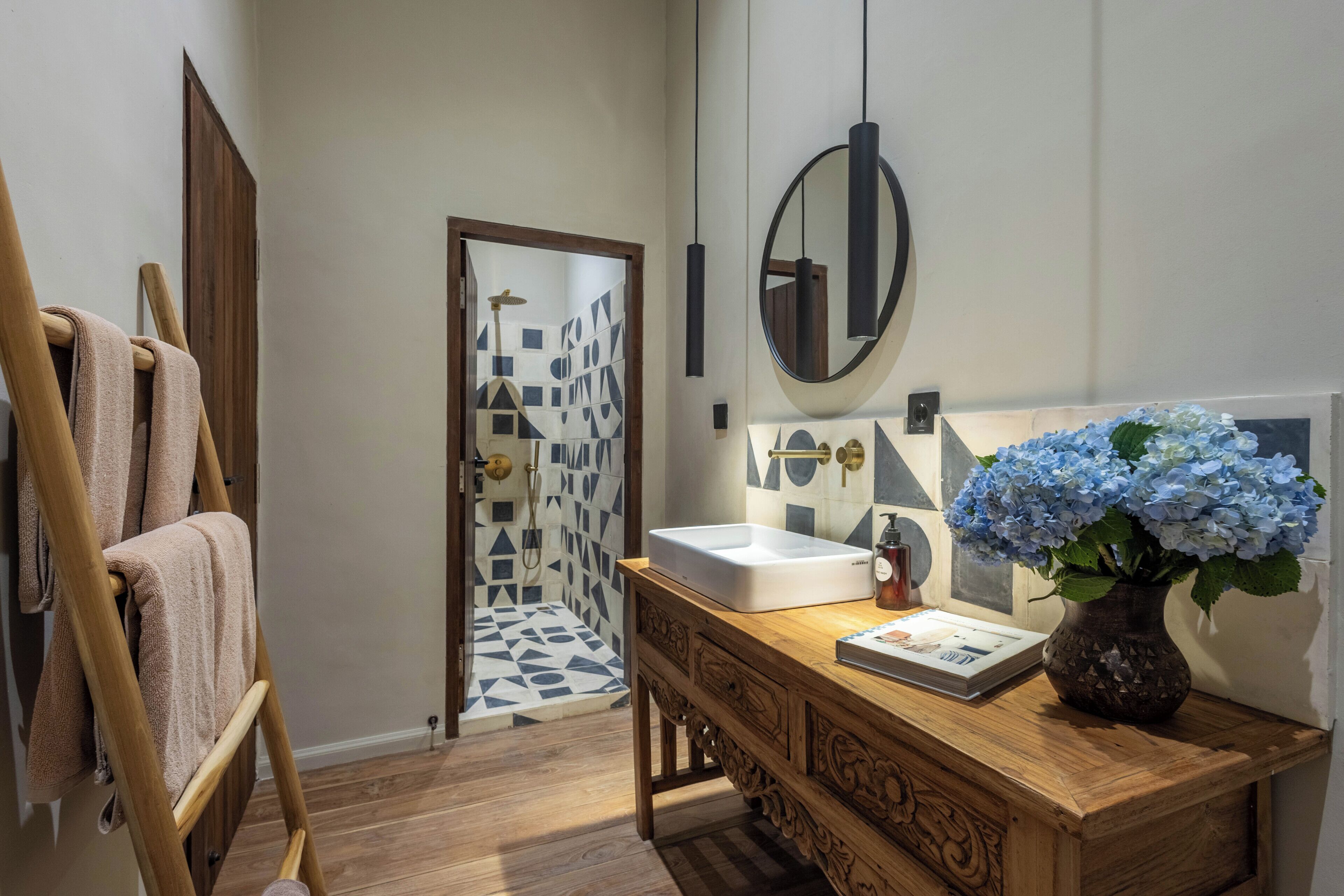 Un baño con tocador de madera, un lavabo blanco, un espejo, un florero con flores y una zona de ducha con diseño geométrico.