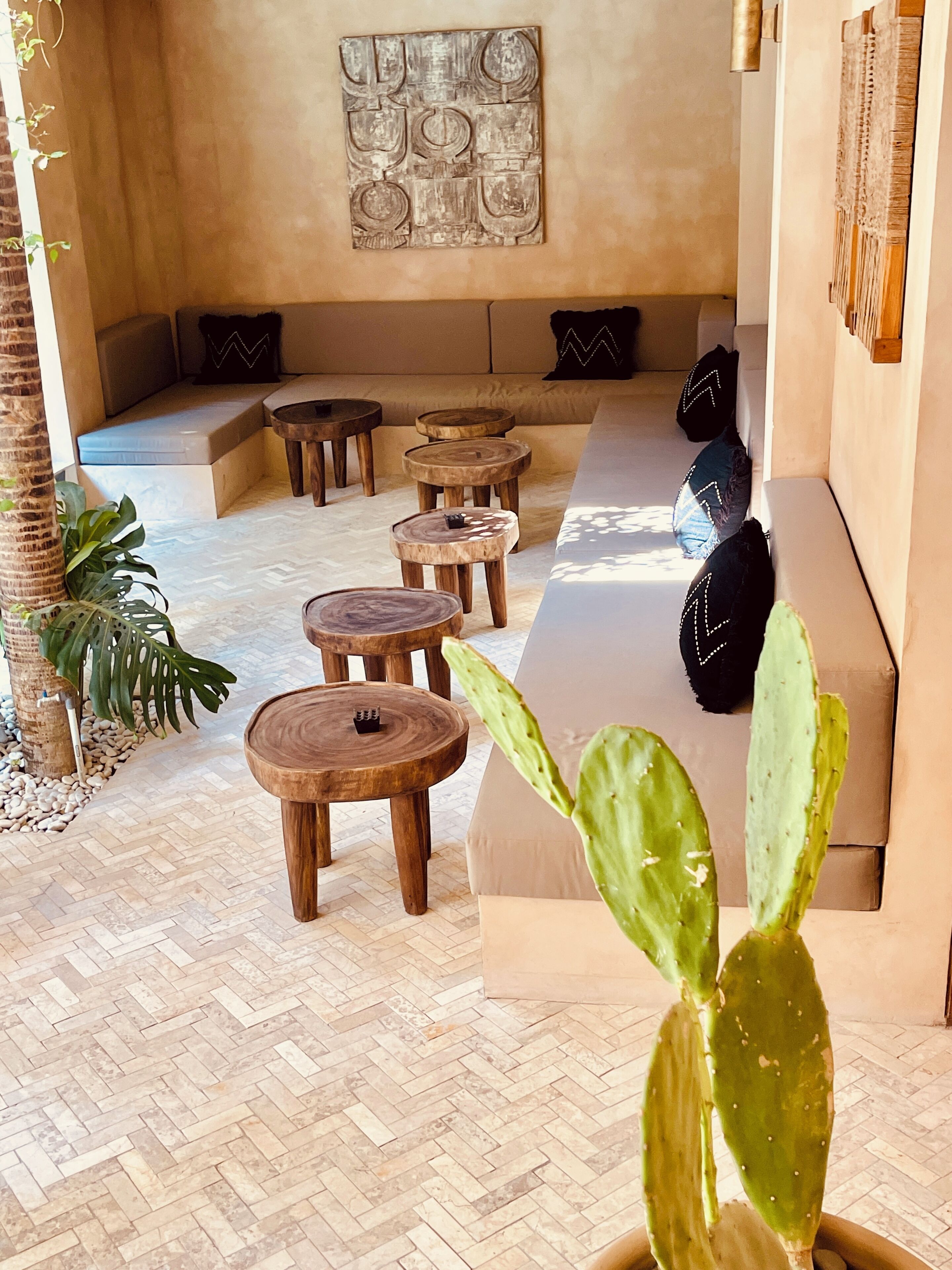 A cozy lounge area with a herringbone floor, wooden stools, a cushioned seating area, and a cactus plant.