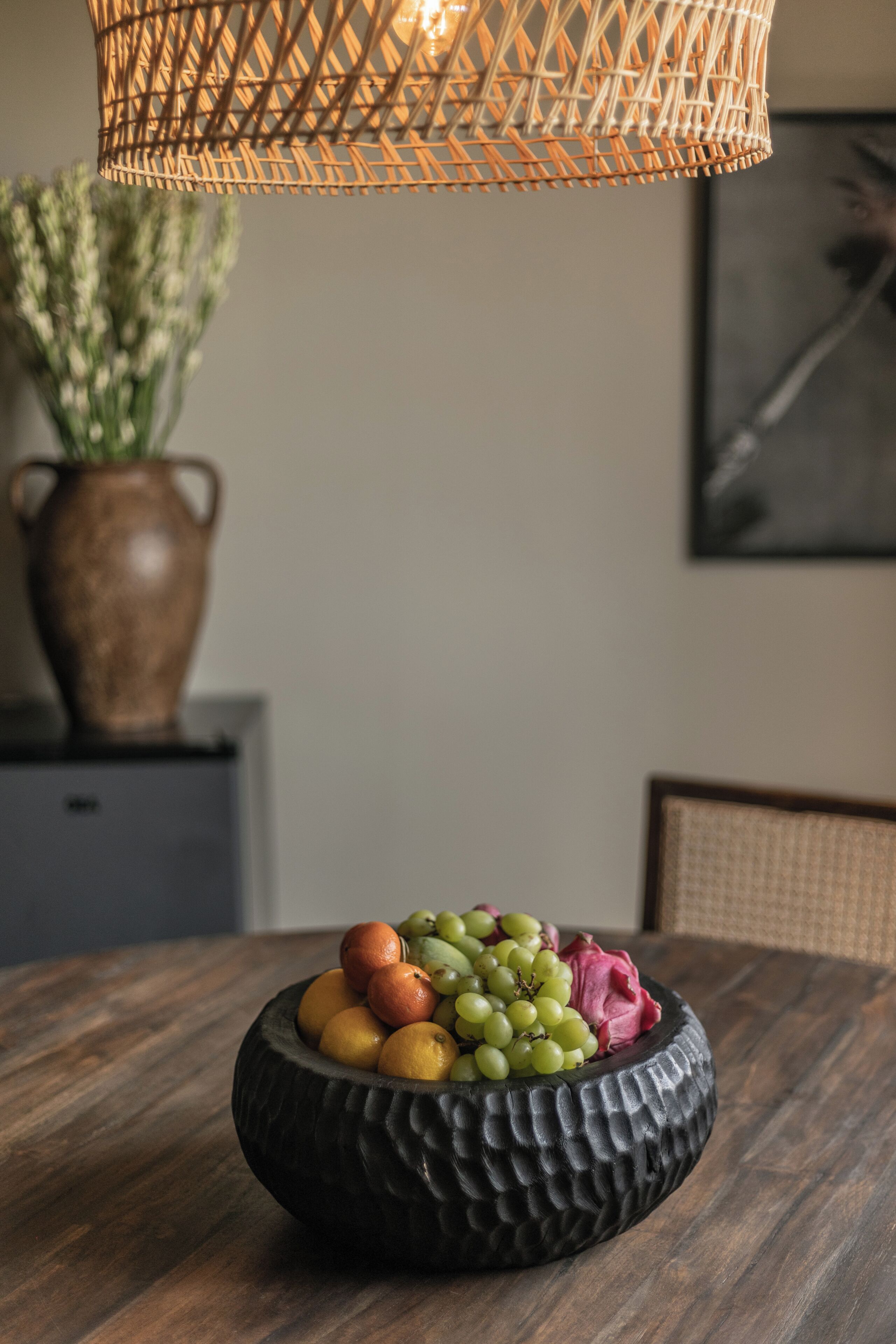 Une table à manger sur laquelle se trouve un saladier texturé rempli de fruits, une lampe en osier et un vase avec des plantes.