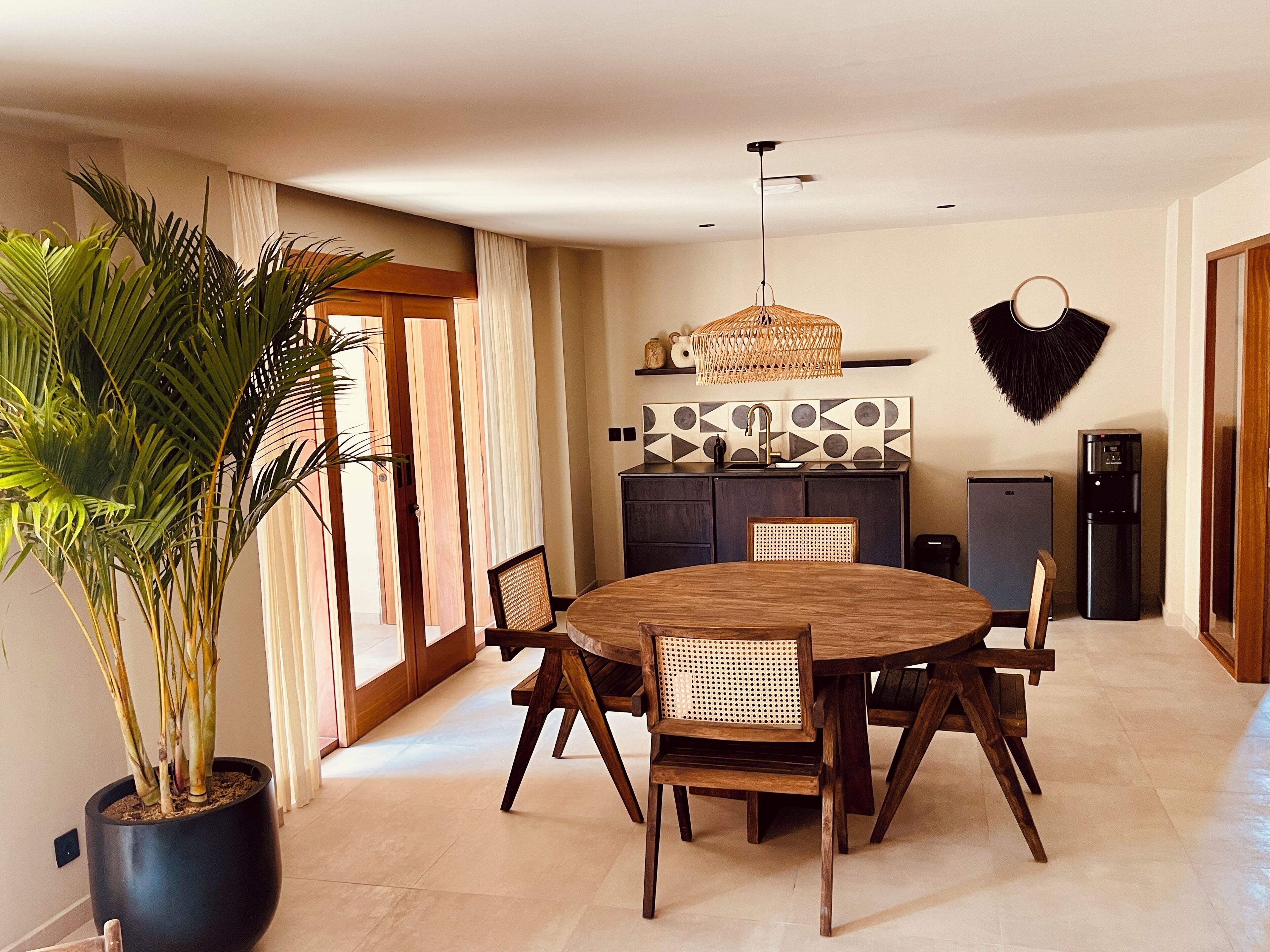 A dining area with a round wooden table and chairs, a potted plant, a kitchen with a geometric backsplash, and a refrigerator.