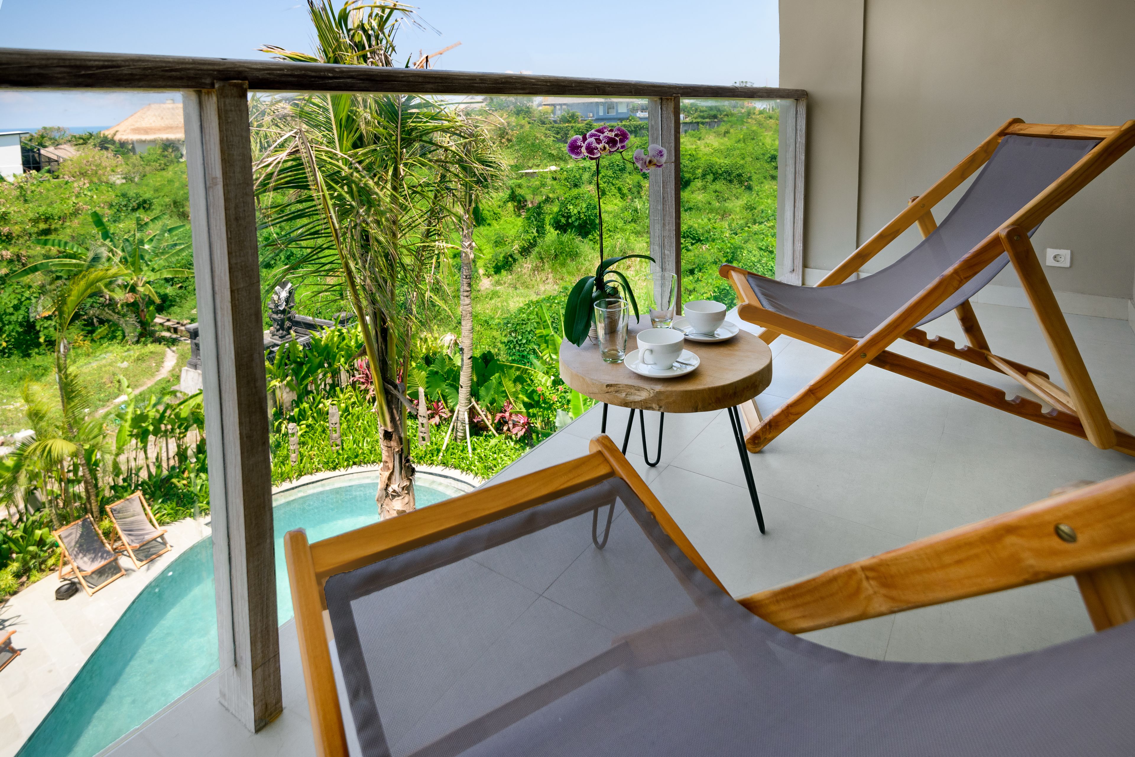 Een balkon met een zwembad, houten ligstoelen, een tafel met bekers en een glas, en uitzicht op weelderige begroeiing en een huis.
